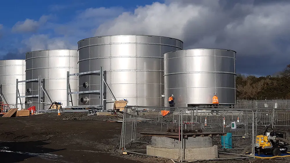 Four large stainless steel tanks with blue sky and clouds in the background a dn two people wearing high vis and hard hats in the foreground
