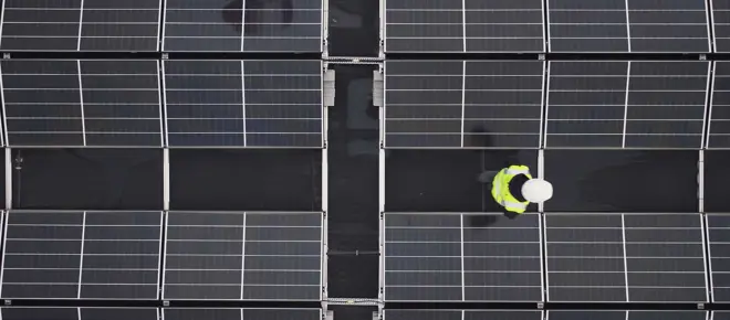 An aerial view of solar panels on a roof with an apprentice engineer in a high visibility jacket and helmet inspecting them.