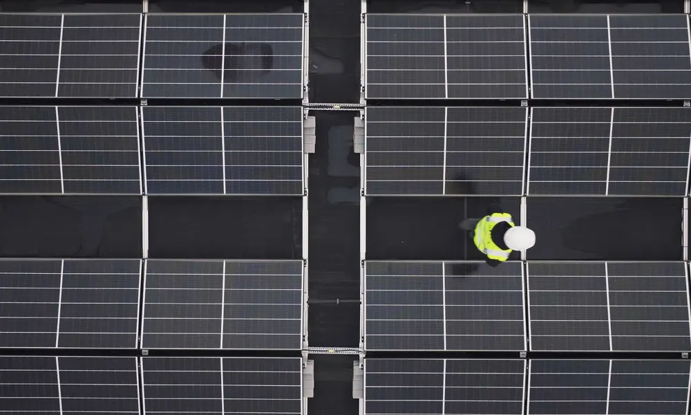 An aerial view of solar panels on a roof with an apprentice engineer in a high visibility jacket and helmet inspecting them.