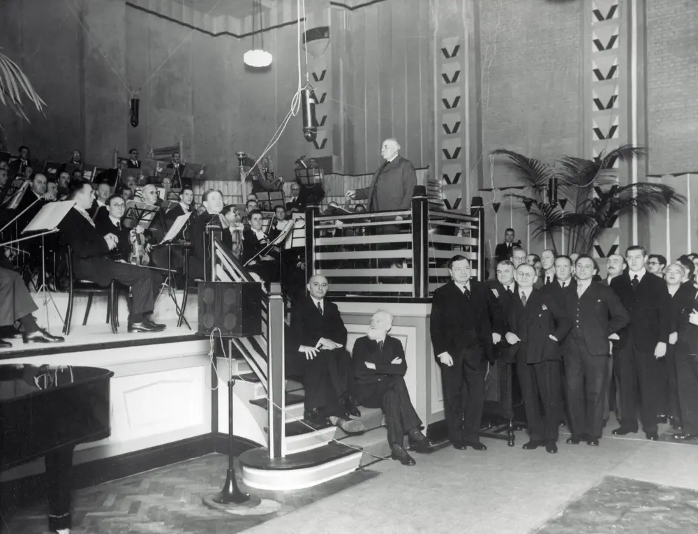 A greyscale image of an orchestra being conducted by Sir Edward Elgar, with a group of people standing near to listen to the acoustics.
