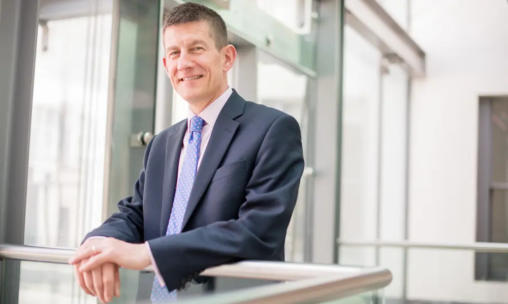A headshot of Dr Shaun Fitzgeralsn FREng wearing a suit, smiling and resting his hands on a banister.