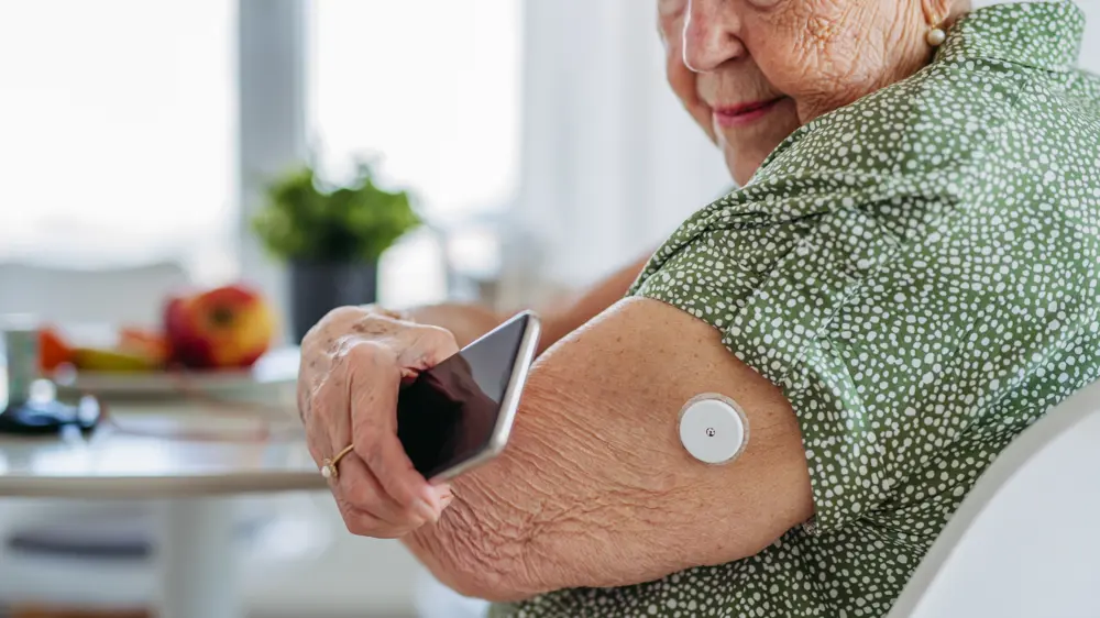 An older white woman wearing a green blouse holds her phone up to her arm, on which she is wearing a continuous glucose monitoring device 