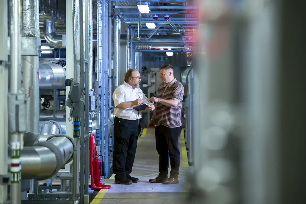 Two men stand and have a conversation in an industrial setting with pipes to the left. One is wearing a white shirt and glasses and holding a pad and pen, while the other is wearing a striped t-shirt and is pointing to something on the pad.  pa