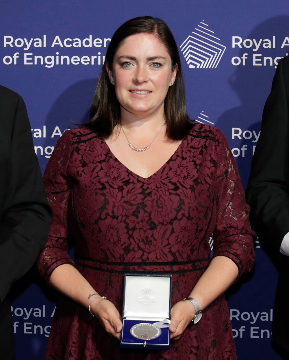 A women with brown hair, wearing a maroon formal dress stands in front of a blue background holding a silver medal in a presentation box