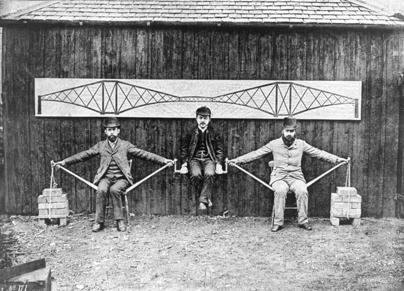 A greyscale image of two men sitting on chairs with their arms open on each side, supporting another man in the middle.