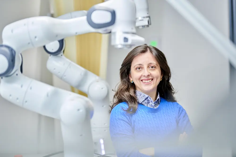 A woman with brown hair and brown eyes smiling at the camera, wearing a blue jumper and standing in front of large robotic arms.