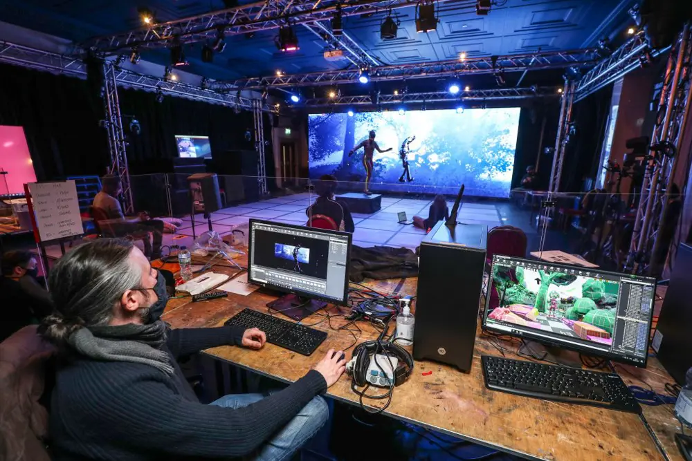 Someone in a motion capture suit in the background, with another person visualising the avatar and fantasy background created on a screen in the foreground.
