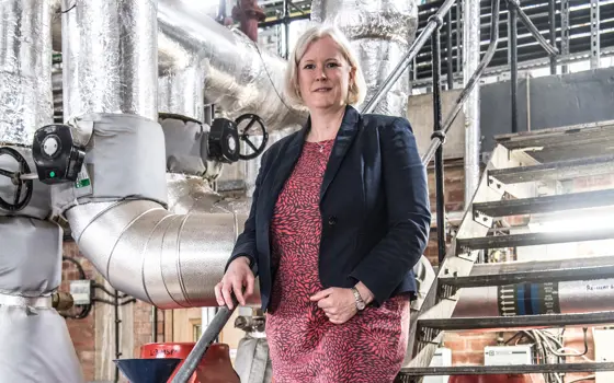 A white woman with blonde hair stand on metal steps with ventilation tubes in the background. She wears a red dress and blue blazer. 
