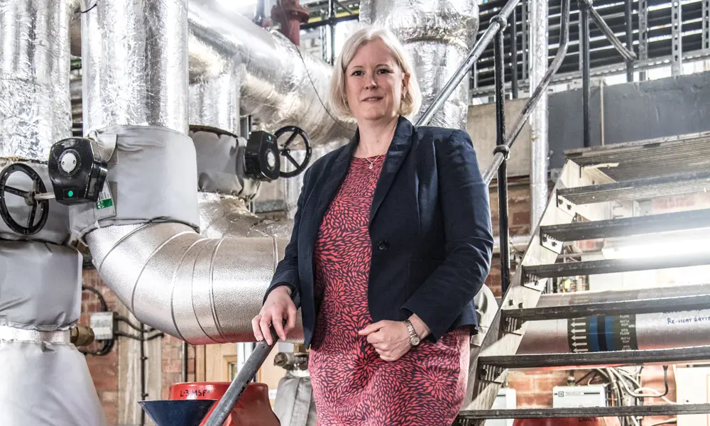 A white woman with blonde hair stand on metal steps with ventilation tubes in the background. She wears a red dress and blue blazer. 