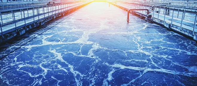 A long tank holds dark wastewater that has light bubbles across its surface