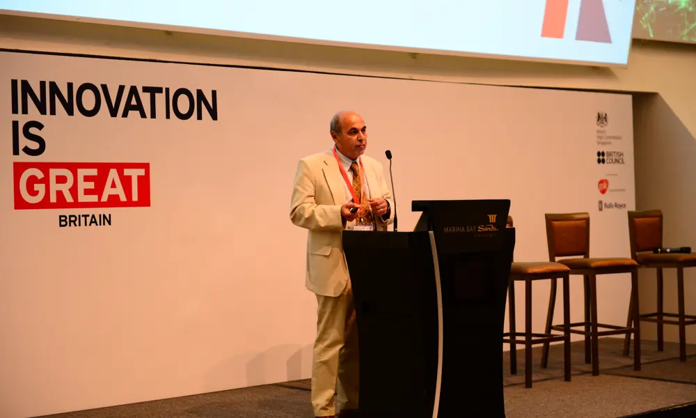 Professor Sir Saeed Zahedi OBE FREng standing on a stage in front of a microphone, giving a talk, with a board saying 'Innovation is great Britain' behind him.