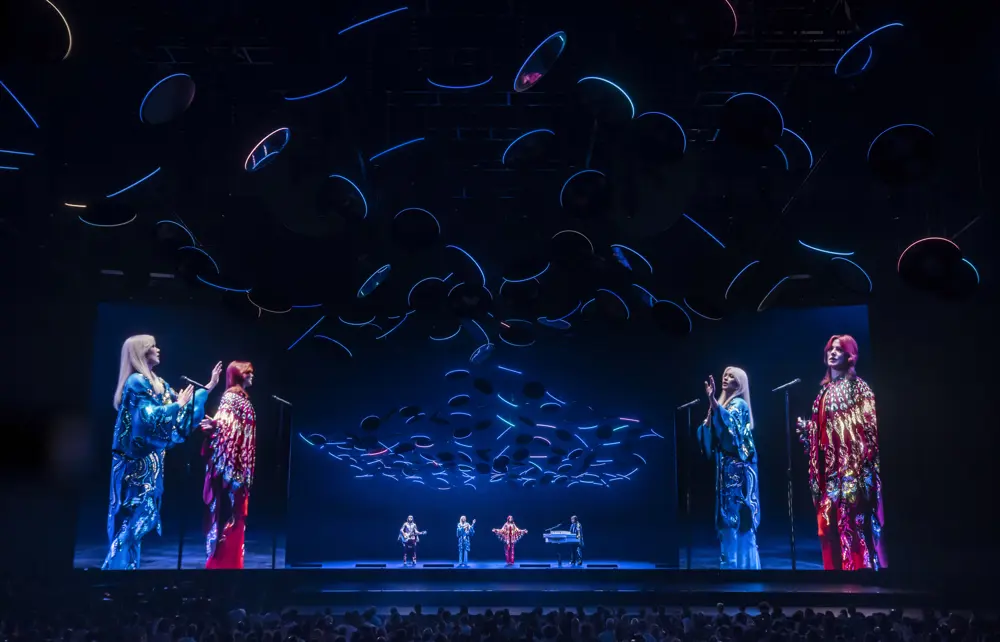 Two singers, a guitarist and a keyboard player wearing elaborately pattered red and blue outfits stand on a stage and perform to a crowd, while subtly illuminated by circular LED lights suspended from the roof
