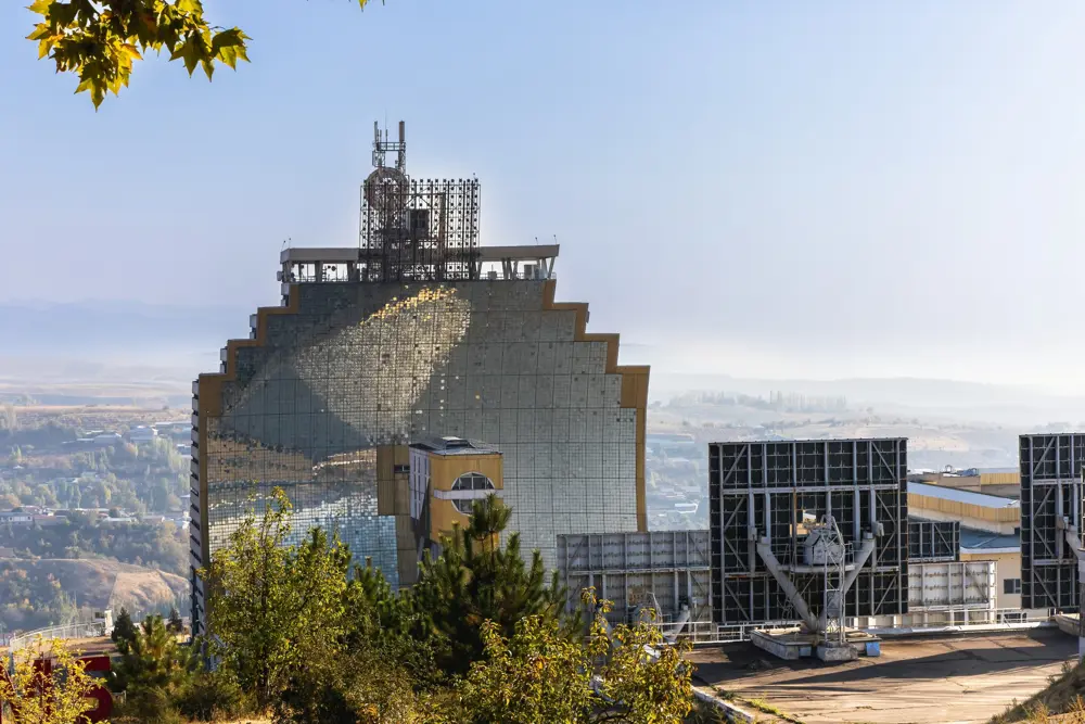 A vast, concave building covered with mirrors glinting in the sun. It is situated on top of the hill with a hazy background of trees, buildings and a blue sky above.