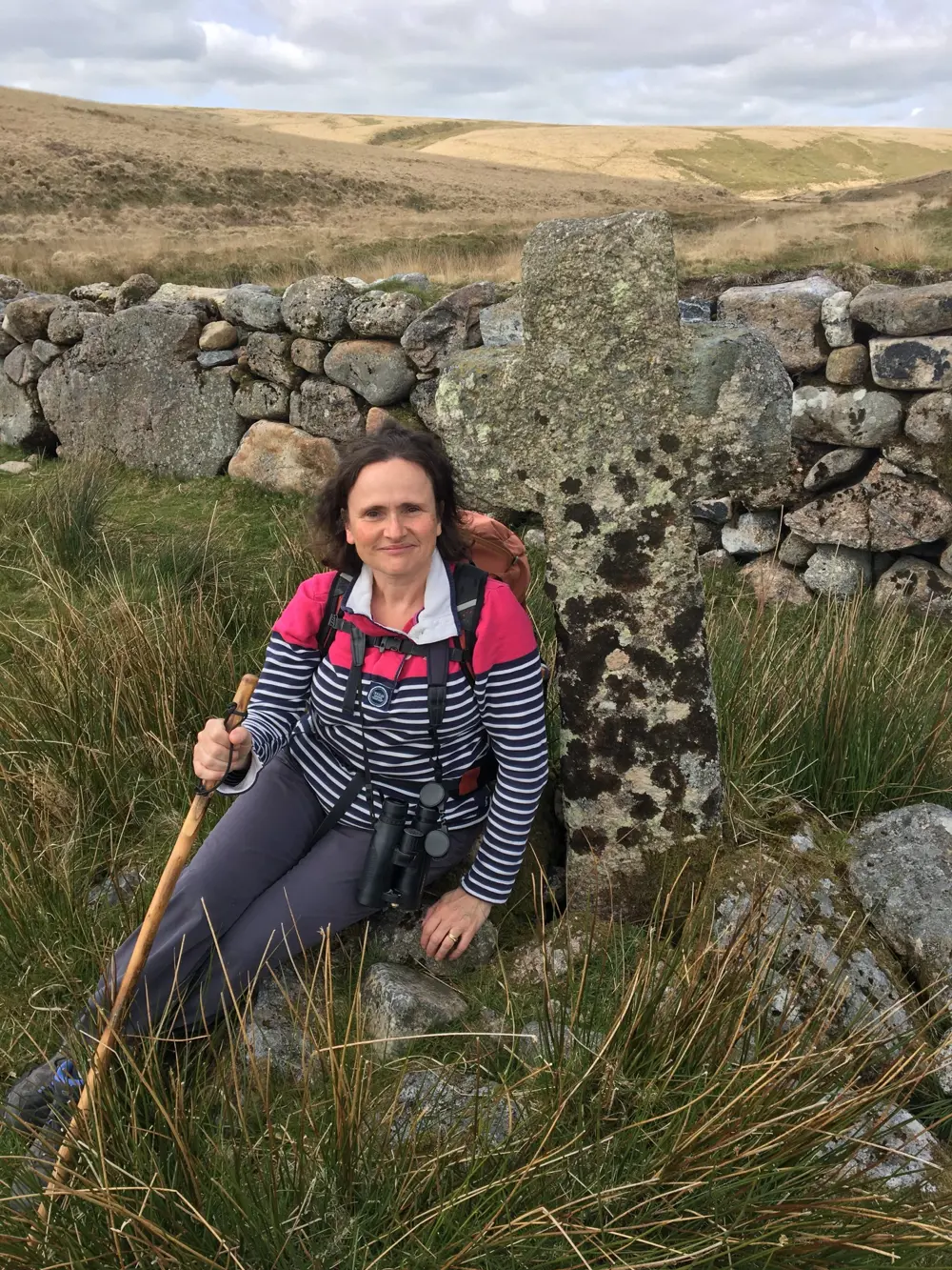 Penny wearing hiking gear with binoculars around her neck, sitting on a rock with moorland in the background.