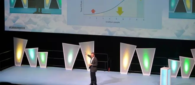 A man onstage in a dark suit, presenting to an audience in front of slides.