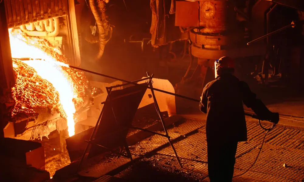 A worker wearing protective gear is standing several metres away from the outlet of a blast furnace, pushing a temperature probe into the white-hot molten metal pouring from it.