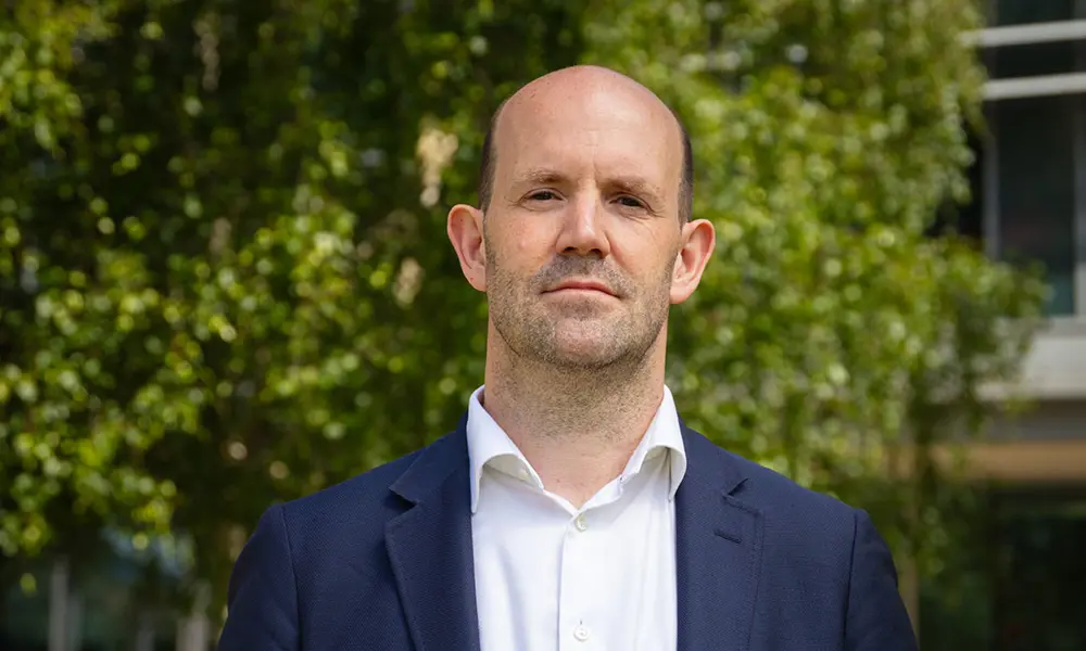 A man looks at the camera. He is wearing a white shirt and a navy blue blazer. He is standing outside with trees behind him