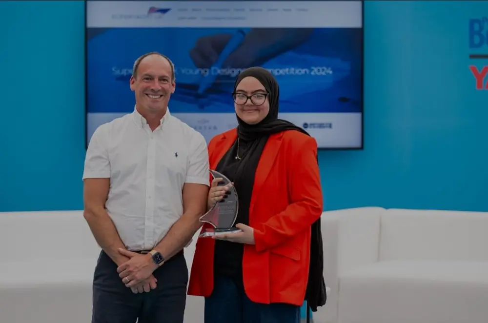 A man in a white shirt stands next to a woman wearing a red jacket, glasses and a headscarf. Both are smiling and looking at the camera. 