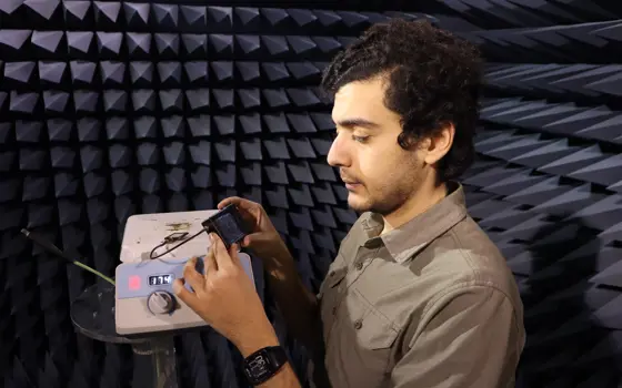 A man with brown hair wearing a beige shirt in a soundproof room, looks at an electronic device held in his hands