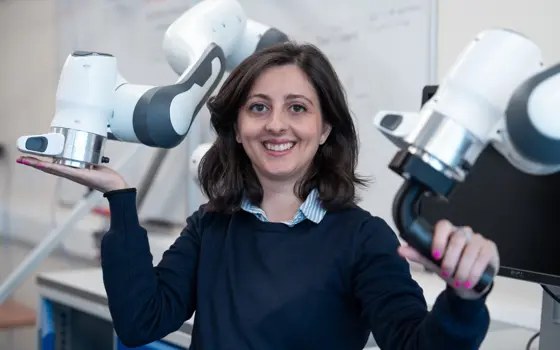 A woman with brown hair and a navy blue jumper posing for a photo, holding a robotic arm with each hand.