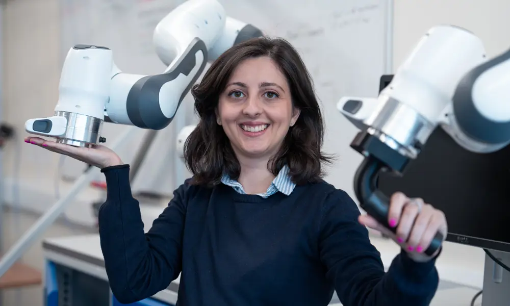 A woman with brown hair and a navy blue jumper posing for a photo, holding a robotic arm with each hand.