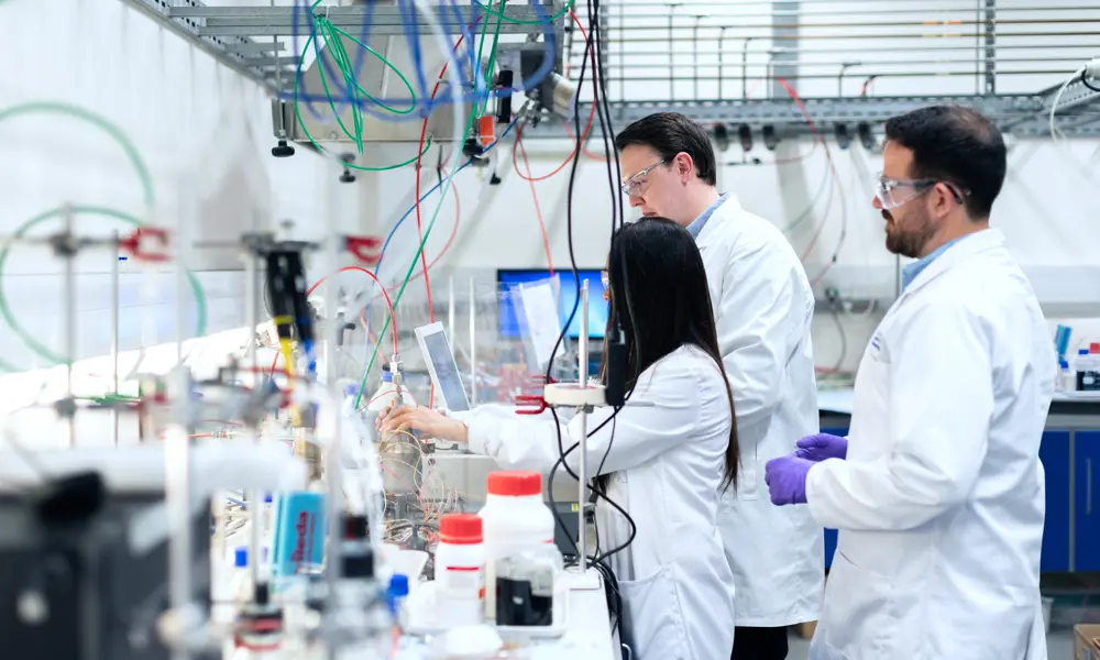 Three engineers in a lab, wearing lab coats and glasses, standing behind a desk full of science equipment, reagents and wires. 