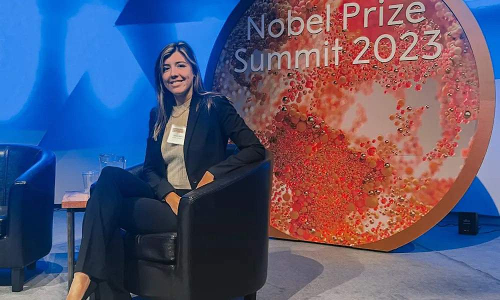 A white woman wearing a black suit sits on a black leather chair on a blue coloured set for a panel talk. Behind her there is a large red circle that says Nobel Prize Summit 2023 in white writing