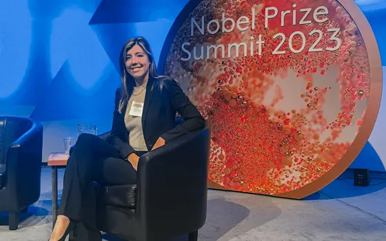 A white woman wearing a black suit sits on a black leather chair on a blue coloured set for a panel talk. Behind her there is a large red circle that says Nobel Prize Summit 2023 in white writing
