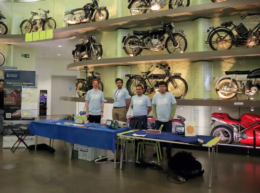Four men wearing light blue t-shirts stand behind two blue tables in front of a wall display that shows a variety of motorbikes