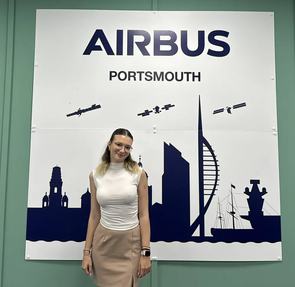A woman wearing glasses, a white top and beige skirt stands in front of a sign saying Airbus Portsmouth