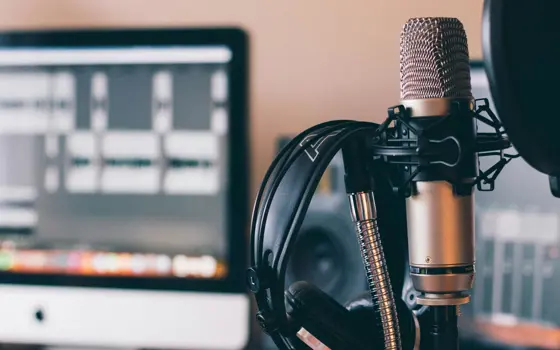 A microphone with headphones and a desk in the background with an iMac computer on it running an audio editing software.