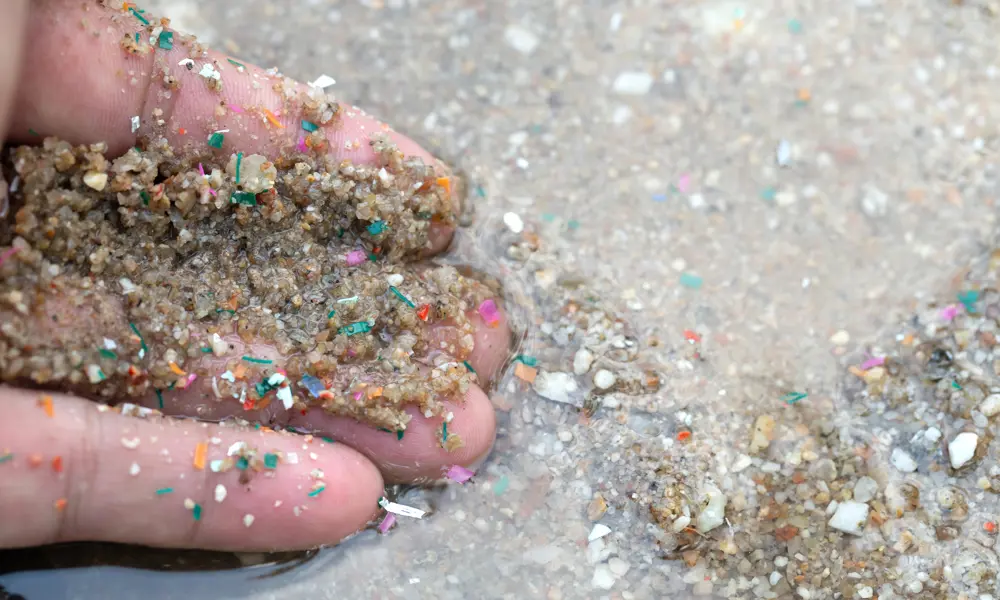 Image shows the fingers from a hand, which is slightly in water having scooped up grains of sand that contain small colourful microplastics