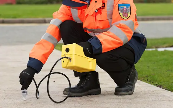 A person in a high visibility jacket, crouching down holding a CableSniffer device, holding a wire to the ground outside.